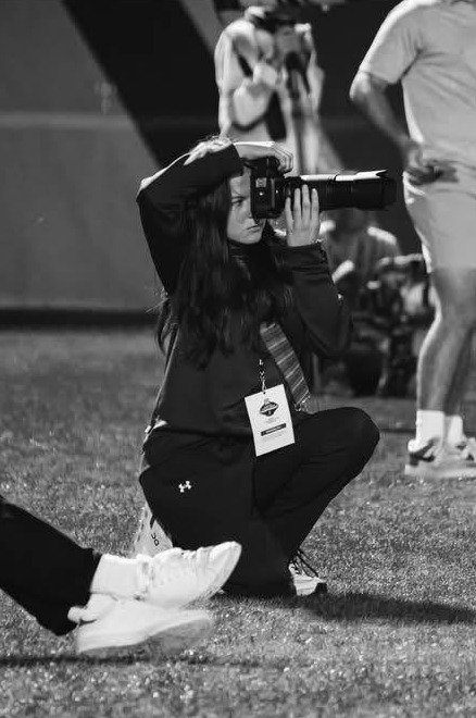 Georgia crouched on a sports sideline at night, photographing the game with a long lens, a press credential around her neck