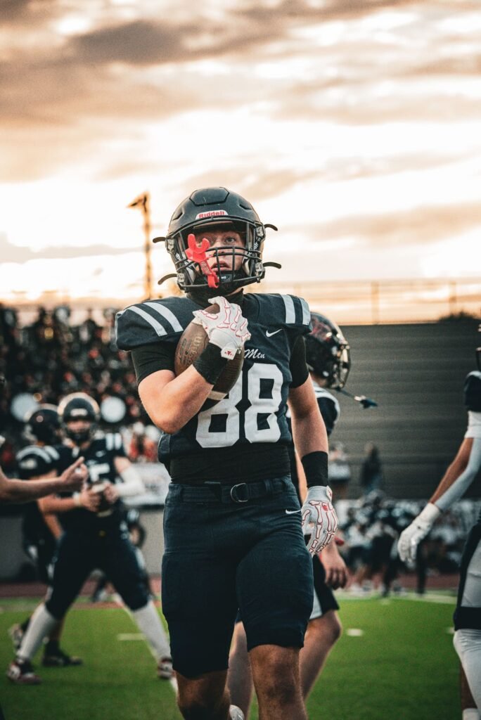 Football player in a dark helmet and number 88 jersey with the sunset behind him, holding the ball