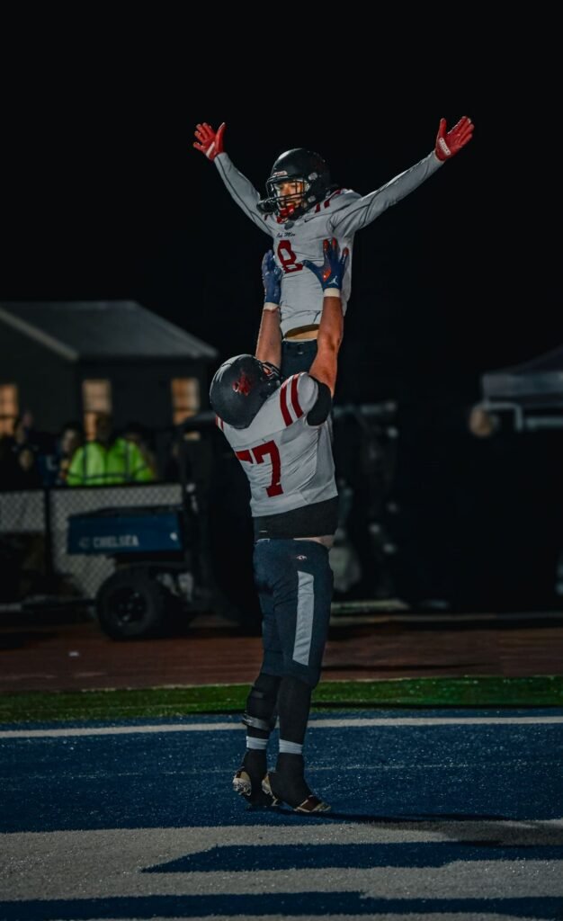 Player lifted onto a teammate's shoulders, arms raised, celebrating on the field