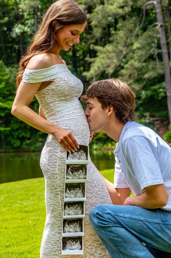 A couple by a pond; she holds a strip of ultrasound prints while he kisses her belly